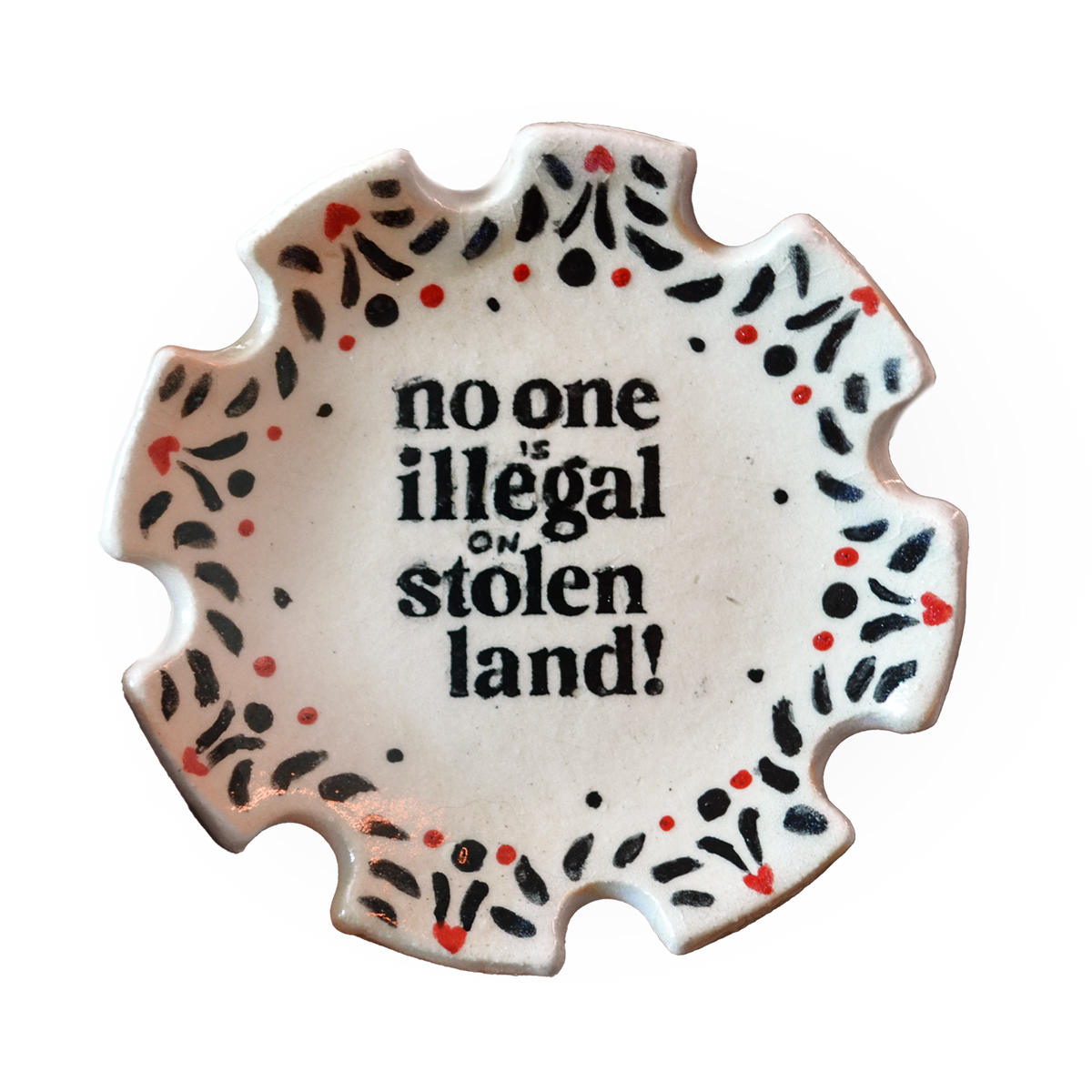 A scalloped ceramic plate with the text 'no one is illegal on stolen land!' in black, surrounded by a decorative border of black and red dots on a white background.