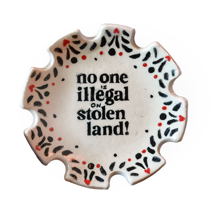 A scalloped ceramic plate with the text 'no one is illegal on stolen land!' in black, surrounded by a decorative border of black and red dots on a white background.