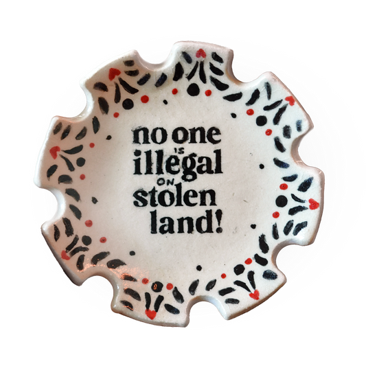 A scalloped ceramic plate with the text 'no one is illegal on stolen land!' in black, surrounded by a decorative border of black and red dots on a white background.
